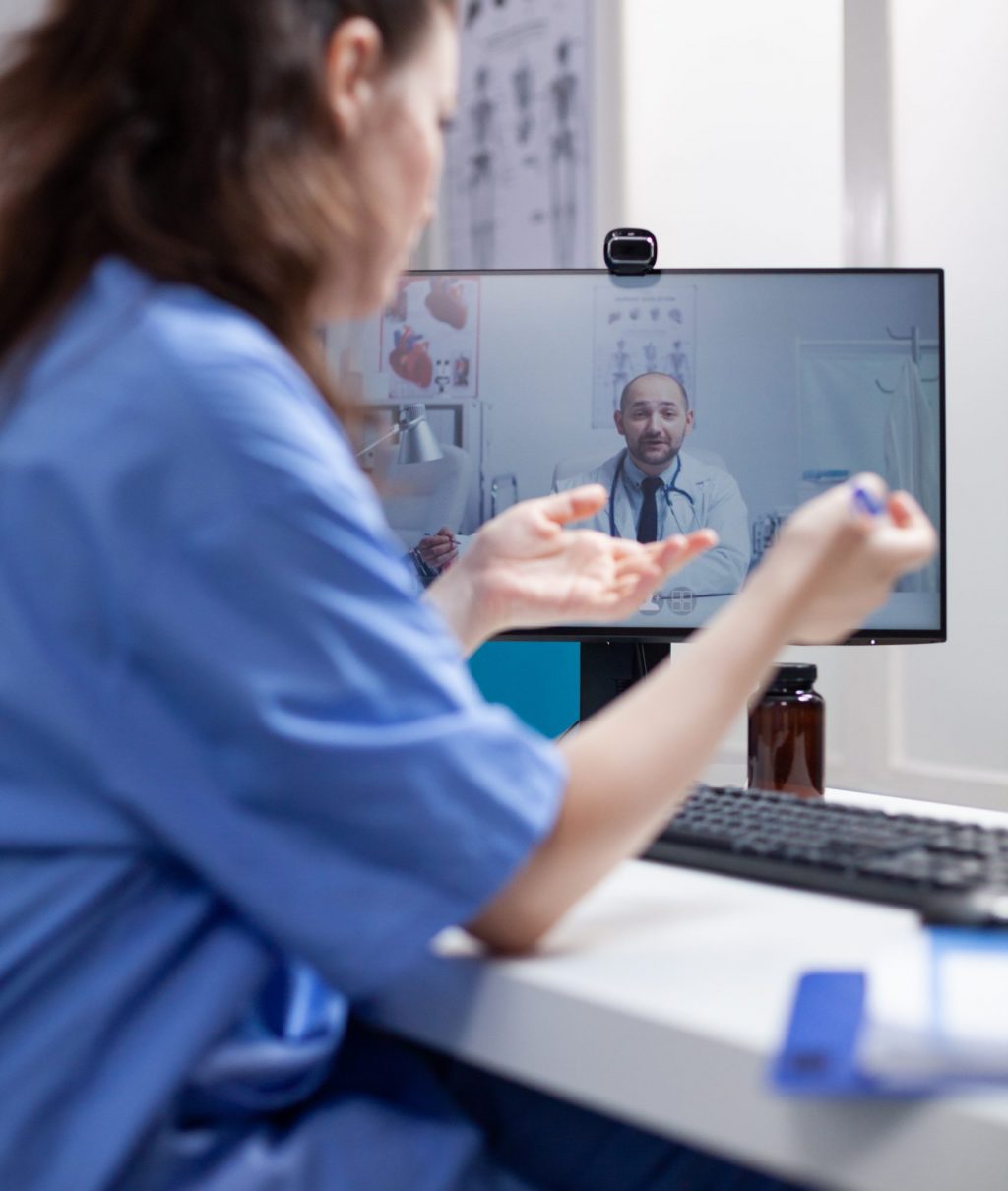 Nurse at desk in telemedicine videocall communicating with doctor specialist in professional medical office. Medic having online internet video conference with young colleague